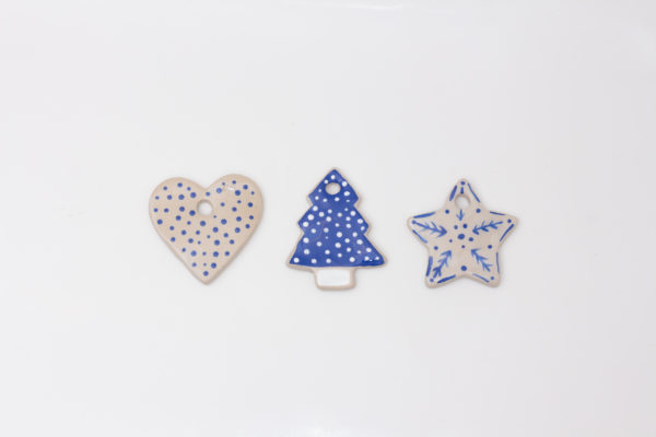 Set of three handmade ceramic ornaments, including a beige heart with blue polka dots, a blue Christmas tree with white dots, and a beige star with blue foliage and dot patterns. Each ornament features a small hole for easy hanging, perfect for festive home decor.