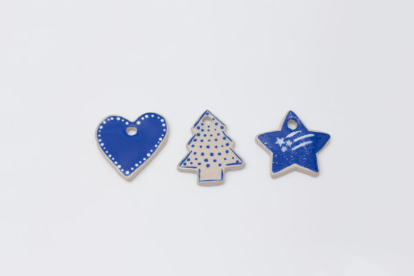 Set of three handmade ceramic ornaments, featuring a blue heart with a white polka-dot border, a beige Christmas tree with blue polka dots and outline, and a blue star with painted white shooting stars and speckled details. Each ornament includes a small hole for easy hanging, ideal for festive holiday decor.